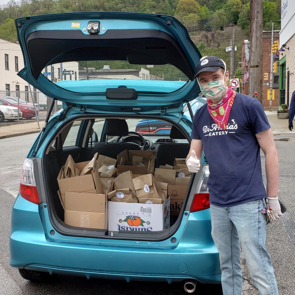 A man with a face mask gives a thumbs up in front of a car loaded with food to be delivered.